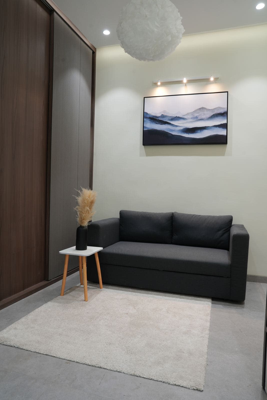 The bedroom set: a dark grey two-seat sofa below a lit framed mountain artwork, a wood-panelled wardrobe alongside and a cloud pendant light above.