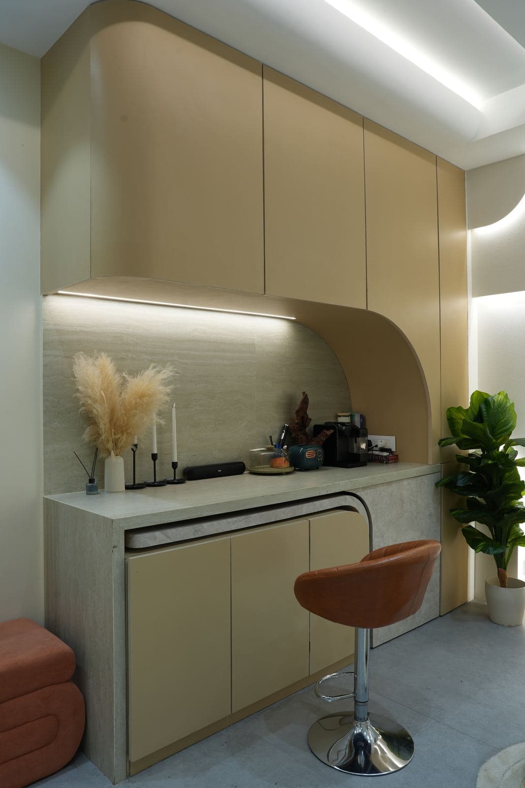 The kitchen set: a curved stone-look counter under lit overhead cabinets, styled with pampas grass, candles and a coffee machine, a tan bar stool at the edge.