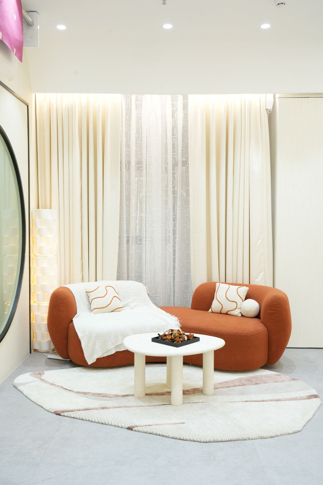 The living room set: the rust-coloured sofa and chaise under cream curtains and a sheer panel, a round white table on the patterned rug, the round mirror door at the left edge.