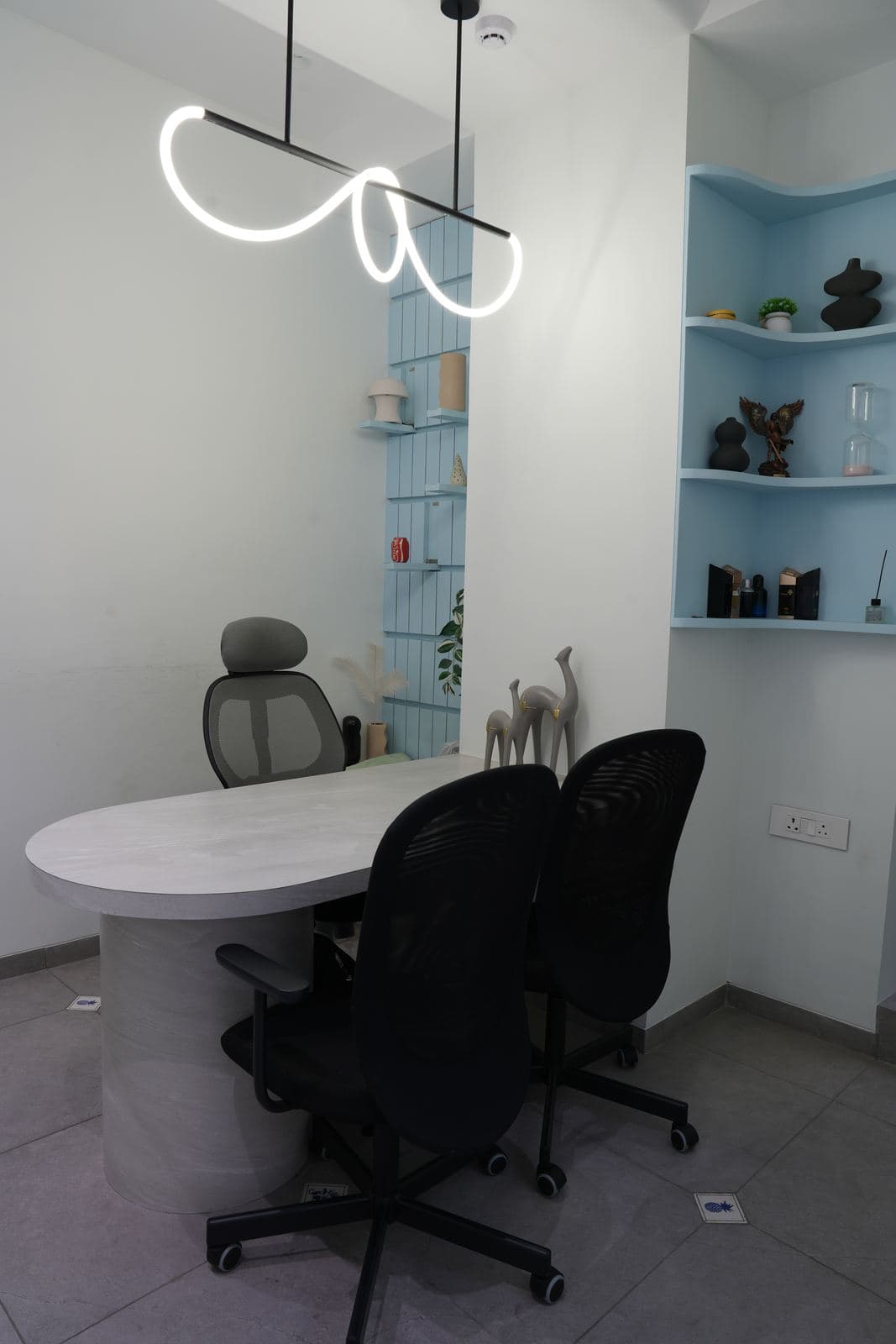 The office desk set: an oval desk and three mesh chairs under a neon squiggle pendant, in front of a blue fluted shelf nook holding small ornaments.